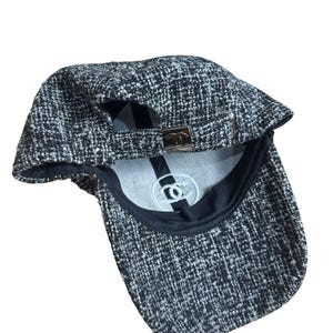 Op de afbeelding: Een zwart-witte tweed baseballcap met een gebogen klep. De cap heeft een metalen sluiting aan de achterkant voor verstelling en een wit geborduurd logo aan de binnenkant. Het ontwerp van de cap is een klassieke stijl.