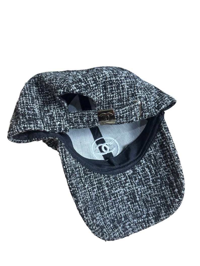 Op de afbeelding: Een zwart-witte tweed baseballcap met een gebogen klep. De cap heeft een metalen sluiting aan de achterkant voor verstelling en een wit geborduurd logo aan de binnenkant. Het ontwerp van de cap is een klassieke stijl.