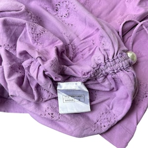 May include: A lavender-colored top with intricate cut-out details and a gathered sleeve. The top features a pearl-like embellishment and a tag with the brand name and product information. The fabric appears soft and lightweight.