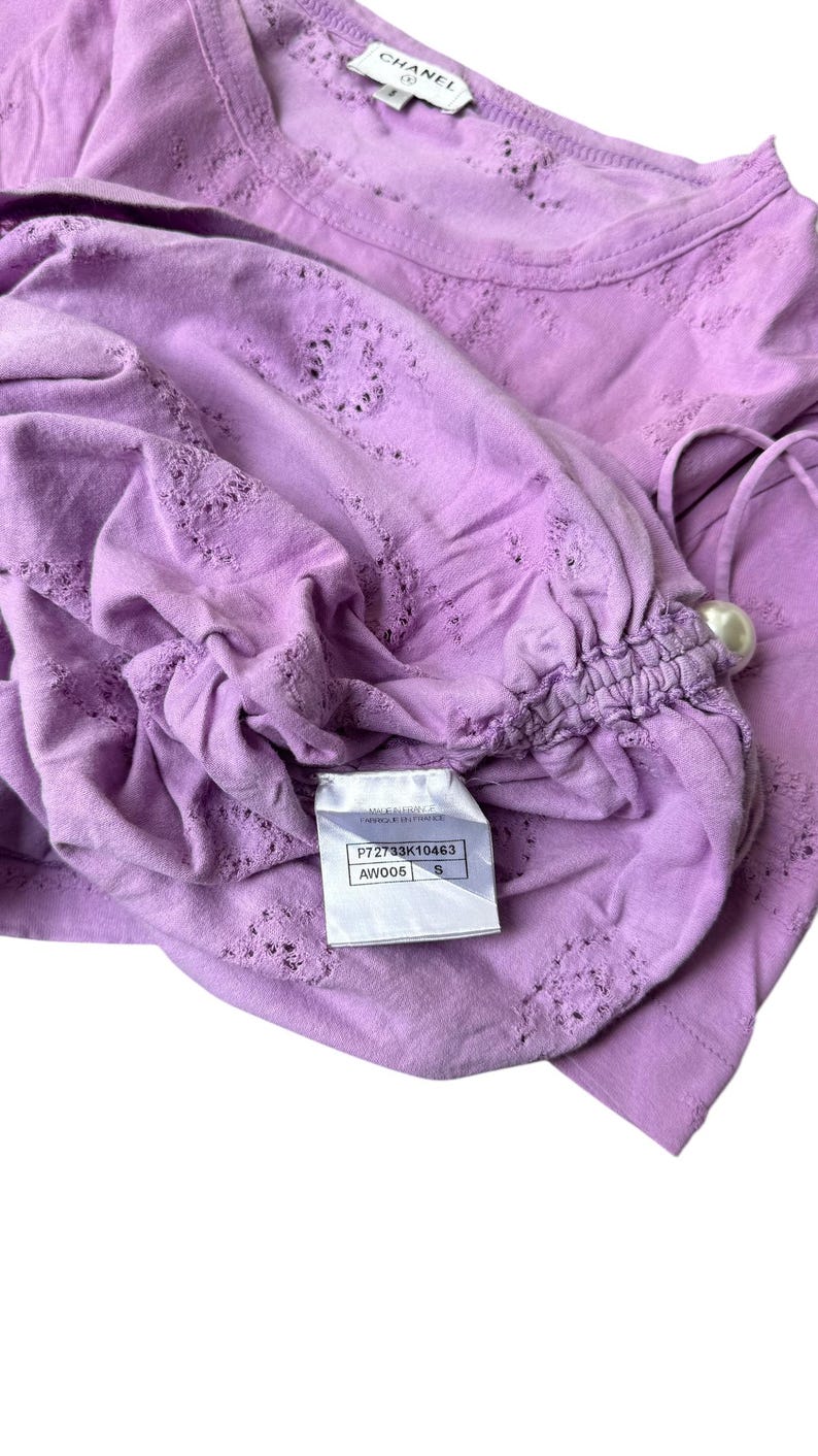 May include: A lavender-colored top with intricate cut-out details and a gathered sleeve. The top features a pearl-like embellishment and a tag with the brand name and product information. The fabric appears soft and lightweight.