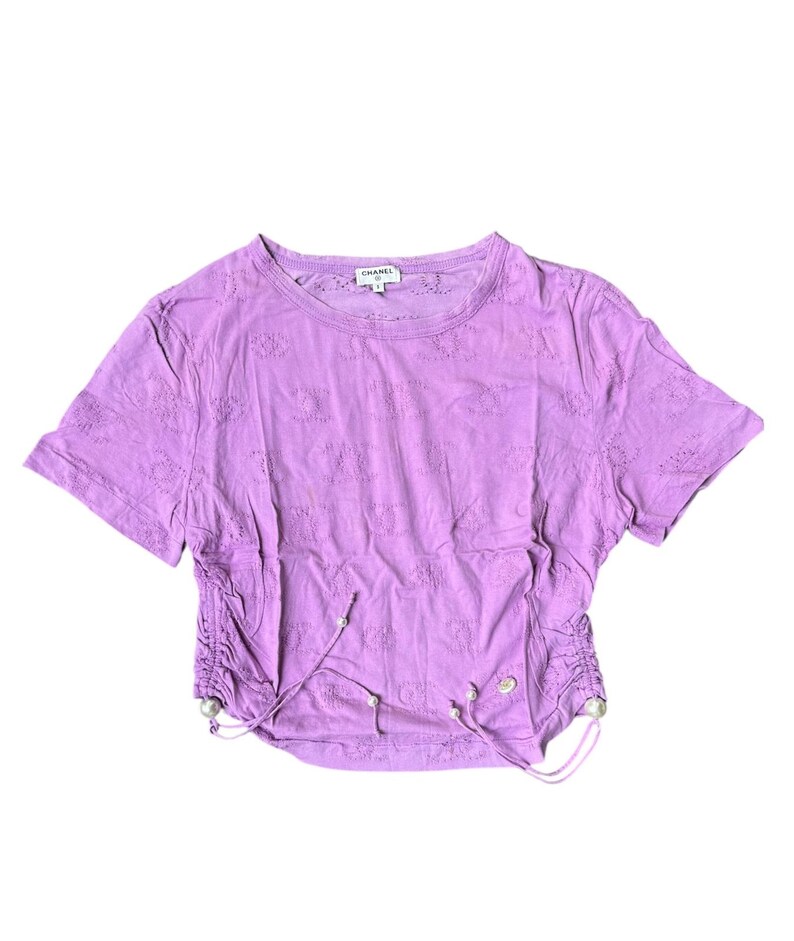 May include: A lavender-colored, short-sleeved crop top with a round neckline. The shirt features an all-over floral eyelet pattern and drawstring details on the sides. The brand name "CHANEL" is visible on the tag.