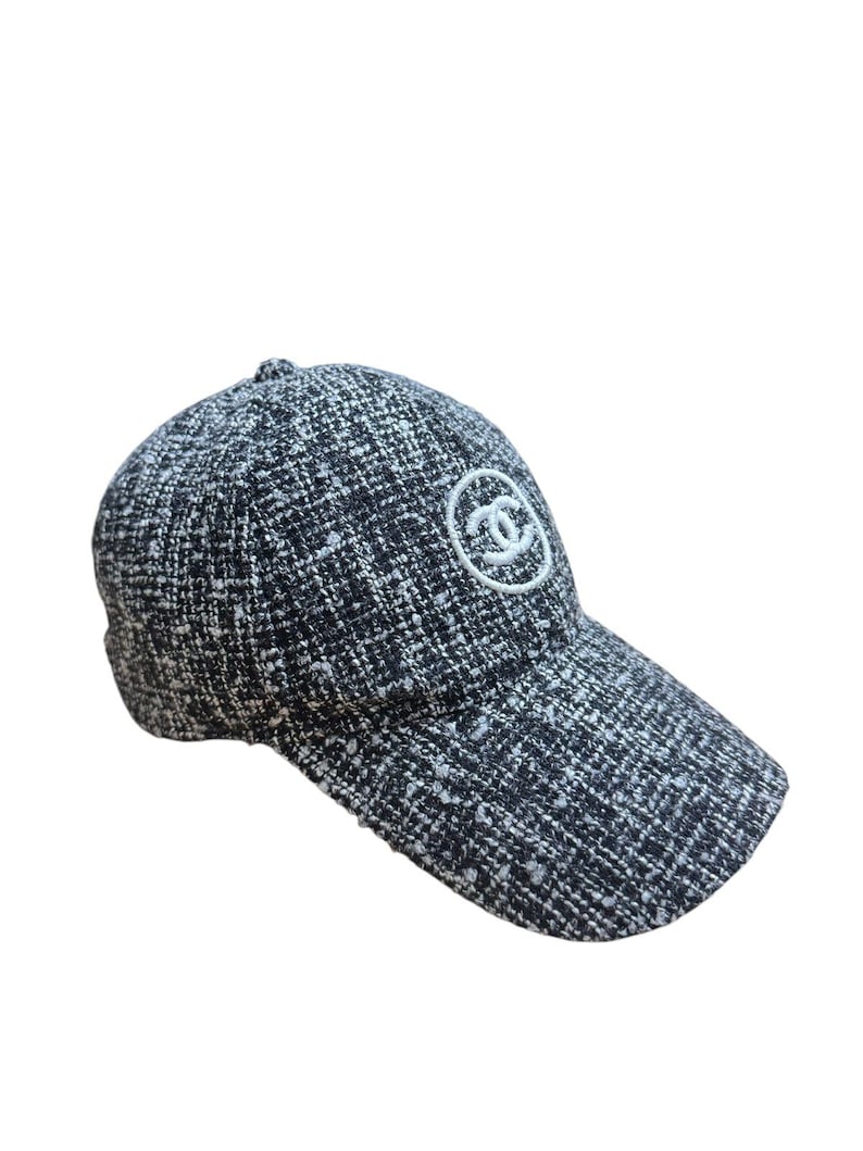 Op de afbeelding: Een zwart-witte tweed baseballcap met een gebogen klep. De pet heeft een wit geborduurd logo op de voorkant. Een klassiek accessoire.