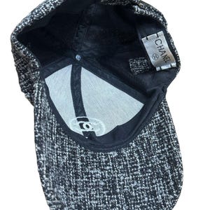 Op de afbeelding: Een zwart-witte tweed baseballcap met een gebogen klep. De binnenkant heeft een witte voering en een label met de tekst "CHANEL". De cap heeft een klassiek zes-panelen ontwerp.