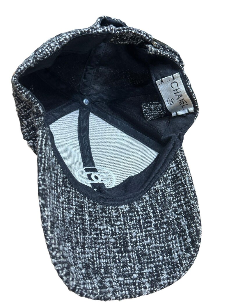 Op de afbeelding: Een zwart-witte tweed baseballcap met een gebogen klep. De binnenkant heeft een witte voering en een label met de tekst "CHANEL". De cap heeft een klassiek zes-panelen ontwerp.