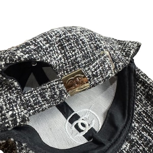 Op de afbeelding: Een zwart-witte tweed baseballcap met een goudkleurige metalen sluiting. De binnenkant heeft een wit geborduurd logo en een zwarte klep. De cap is gemaakt van een textuurstof.