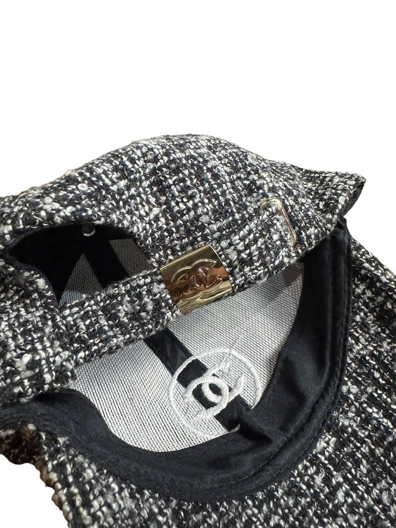 Op de afbeelding: Een zwart-witte tweed baseballcap met een goudkleurige metalen sluiting. De binnenkant heeft een wit geborduurd logo en een zwarte klep. De cap is gemaakt van een textuurstof.