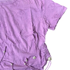 May include: A lilac-colored t-shirt with a crew neck and short sleeves. The shirt features a textured pattern and drawstring details with faux pearl accents. A small logo is visible on the upper left. The shirt is made of a soft material.