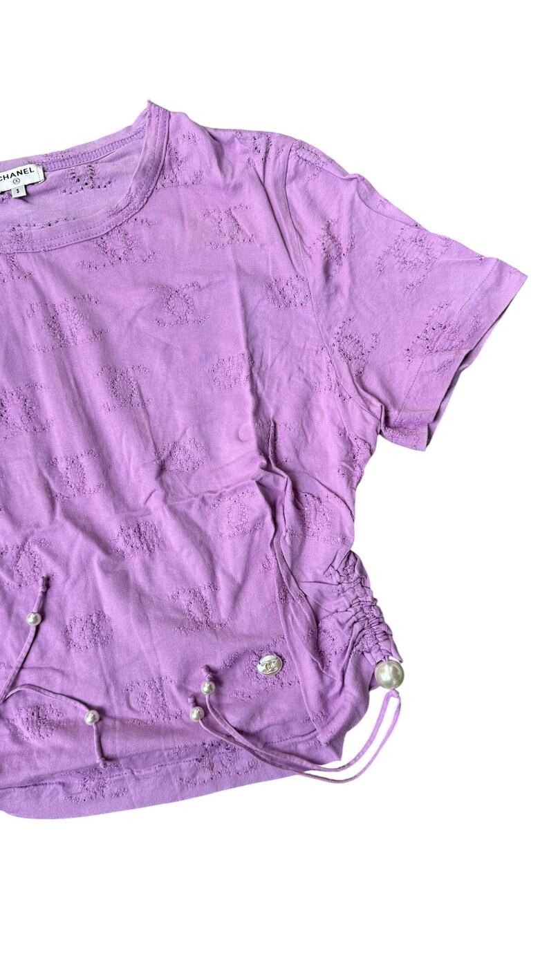 May include: A lilac-colored t-shirt with a crew neck and short sleeves. The shirt features a textured pattern and drawstring details with faux pearl accents. A small logo is visible on the upper left. The shirt is made of a soft material.