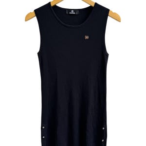 May include: A sleeveless black ribbed knit top featuring a round neckline and a small square logo patch on the upper left chest. The garment includes three small silver-toned buttons on each side of the hem and a Chanel brand label at the neck.
