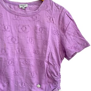 May include: Lilac-colored short-sleeved shirt with a round neckline and a decorative pattern. The shirt has a drawstring detail on the side and a small logo near the neckline. The shirt is hanging on a wooden hanger.