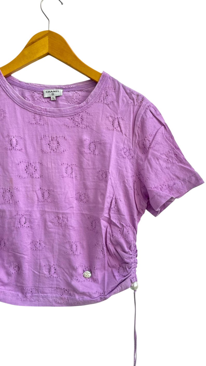 May include: Lilac-colored short-sleeved shirt with a round neckline and a decorative pattern. The shirt has a drawstring detail on the side and a small logo near the neckline. The shirt is hanging on a wooden hanger.