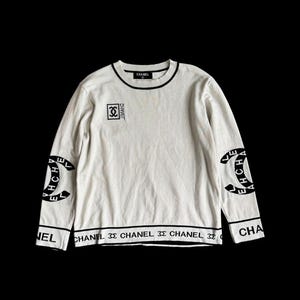 May include: Cream-coloured sweater with black trim and the word "CHANEL" printed on it. The sweater features the Chanel logo on the sleeves and chest. The word "CHANEL" is also printed on the bottom of the sweater.