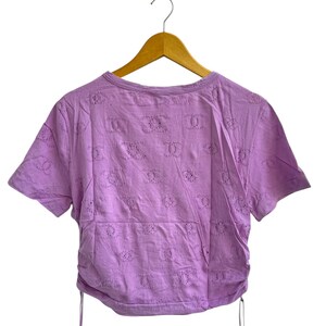 May include: A short-sleeved, lilac-colored top with a round neckline and eyelet detailing. The shirt has a drawstring at the hem and is hanging on a wooden hanger against a white background. The eyelet pattern is repeated across the fabric.