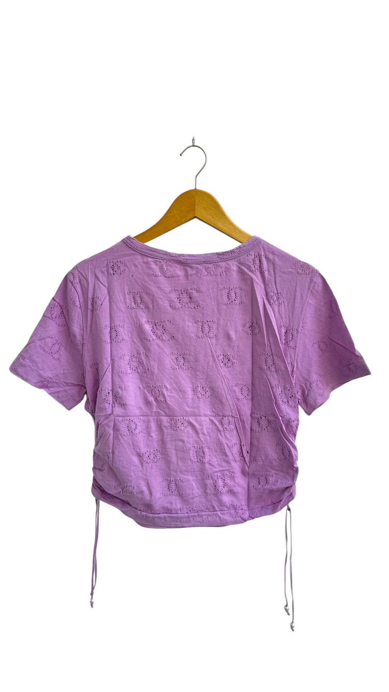 May include: A short-sleeved, lilac-colored top with a round neckline and eyelet detailing. The shirt has a drawstring at the hem and is hanging on a wooden hanger against a white background. The eyelet pattern is repeated across the fabric.