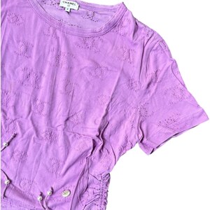 May include: A lilac-colored short-sleeved t-shirt with a round neckline. The shirt features a textured pattern and a drawstring detail with faux pearls. The brand name "CHANEL" is visible on the label.