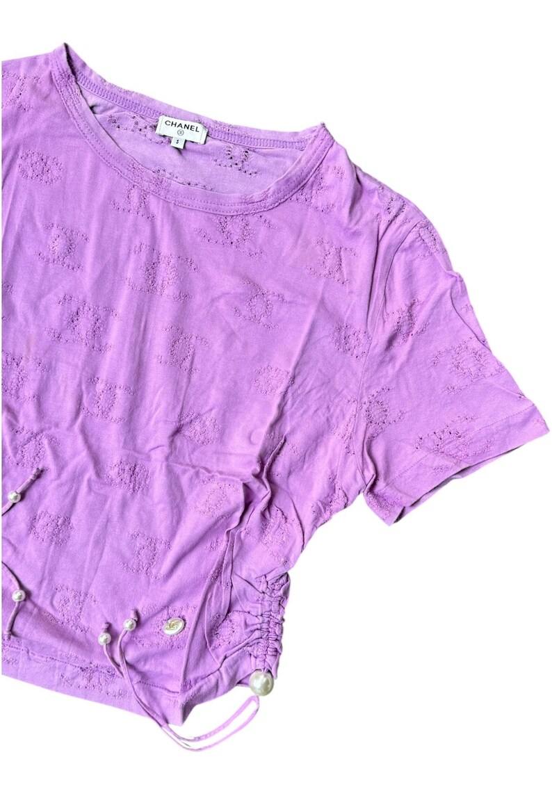 May include: A lilac-colored short-sleeved t-shirt with a round neckline. The shirt features a textured pattern and a drawstring detail with faux pearls. The brand name "CHANEL" is visible on the label.
