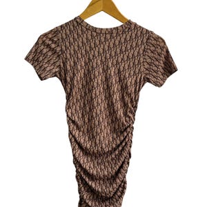 Peut inclure: Haut marron à manches courtes avec un motif répétitif. Le tissu est froncé sur la moitié inférieure. Le haut est suspendu à un cintre en bois sur fond blanc. Le motif semble être un logo.