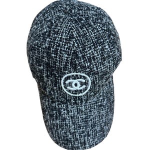 Op de afbeelding: Een zwart-witte tweed baseballcap met een gebogen klep. De cap heeft een wit geborduurd logo op de voorkant. De stof heeft een getextureerde uitstraling.