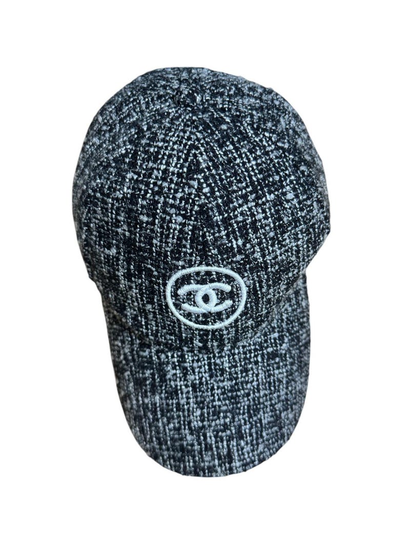 Op de afbeelding: Een zwart-witte tweed baseballcap met een gebogen klep. De cap heeft een wit geborduurd logo op de voorkant. De stof heeft een getextureerde uitstraling.