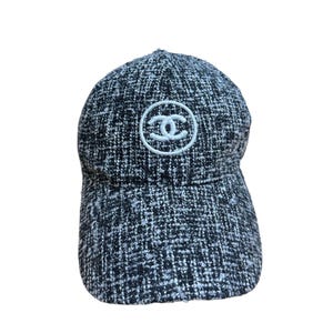 Op de afbeelding: Een zwart-witte tweed baseballcap met een gebogen klep. De cap heeft een wit cirkelvormig embleem met een dubbel C-logo op de voorkant. De hoed is gemaakt van een textuurstof.