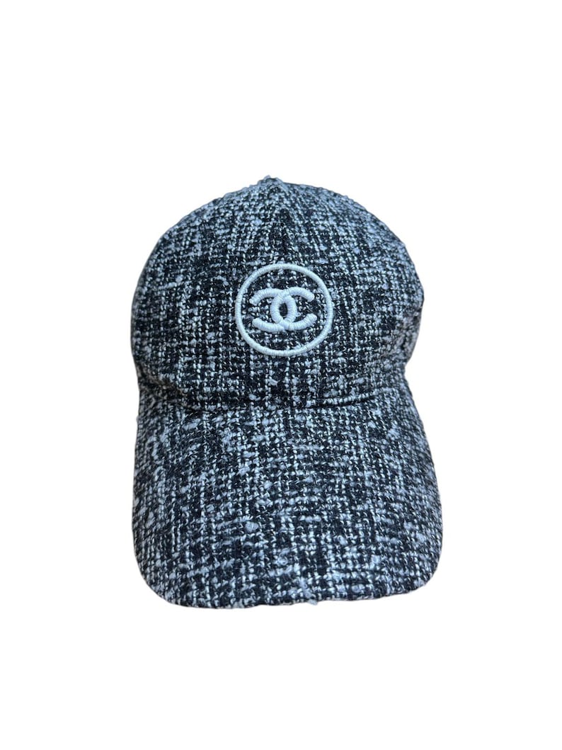 Op de afbeelding: Een zwart-witte tweed baseballcap met een gebogen klep. De cap heeft een wit cirkelvormig embleem met een dubbel C-logo op de voorkant. De hoed is gemaakt van een textuurstof.