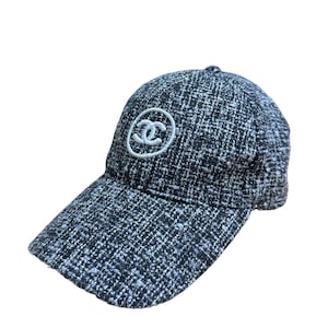 Op de afbeelding: Een zwart-witte tweed baseballcap met een gebogen klep. De pet heeft een wit cirkelvormig logo op de voorkant. De hoed is gemaakt van een geweven stof.