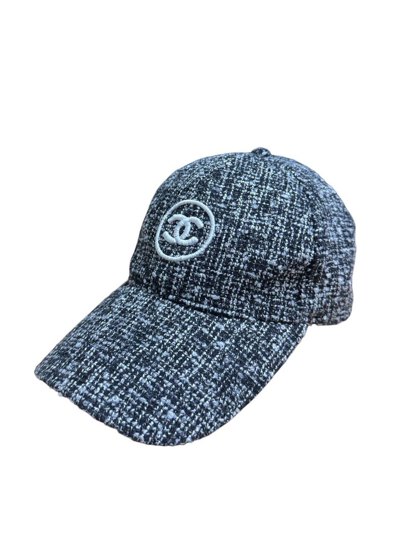 Op de afbeelding: Een zwart-witte tweed baseballcap met een gebogen klep. De pet heeft een wit cirkelvormig logo op de voorkant. De hoed is gemaakt van een geweven stof.