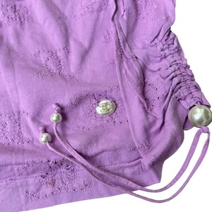 May include: A lilac-colored top with a textured design and drawstring detail. The top features a decorative button and pearl accents. The fabric has a distressed look. The garment is a stylish addition to any wardrobe.