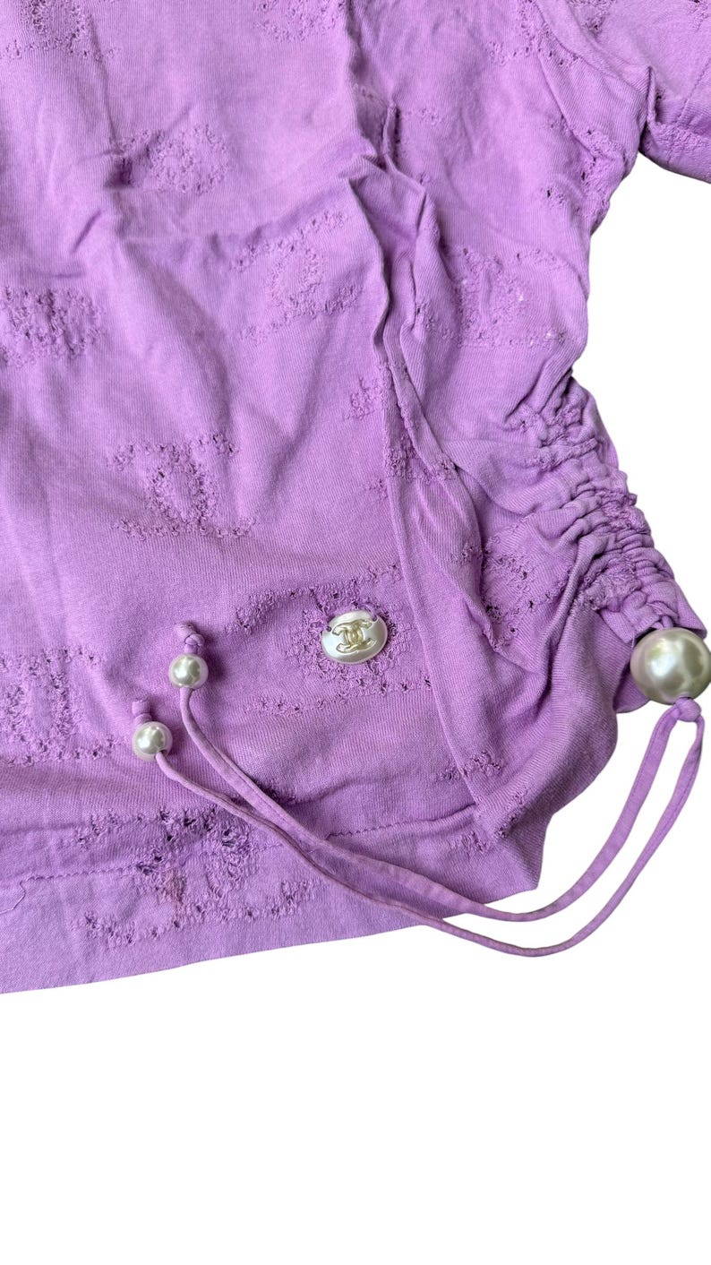 May include: A lilac-colored top with a textured design and drawstring detail. The top features a decorative button and pearl accents. The fabric has a distressed look. The garment is a stylish addition to any wardrobe.