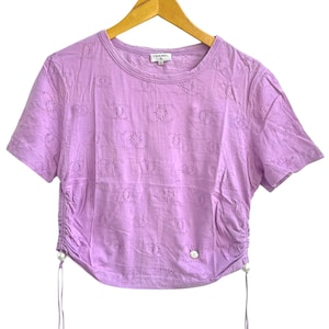 May include: A lavender short-sleeved t-shirt with a crew neck and a drawstring detail on the side. The shirt has a pattern of small, repeating shapes and a small logo near the neckline. The shirt is hanging on a wooden hanger.