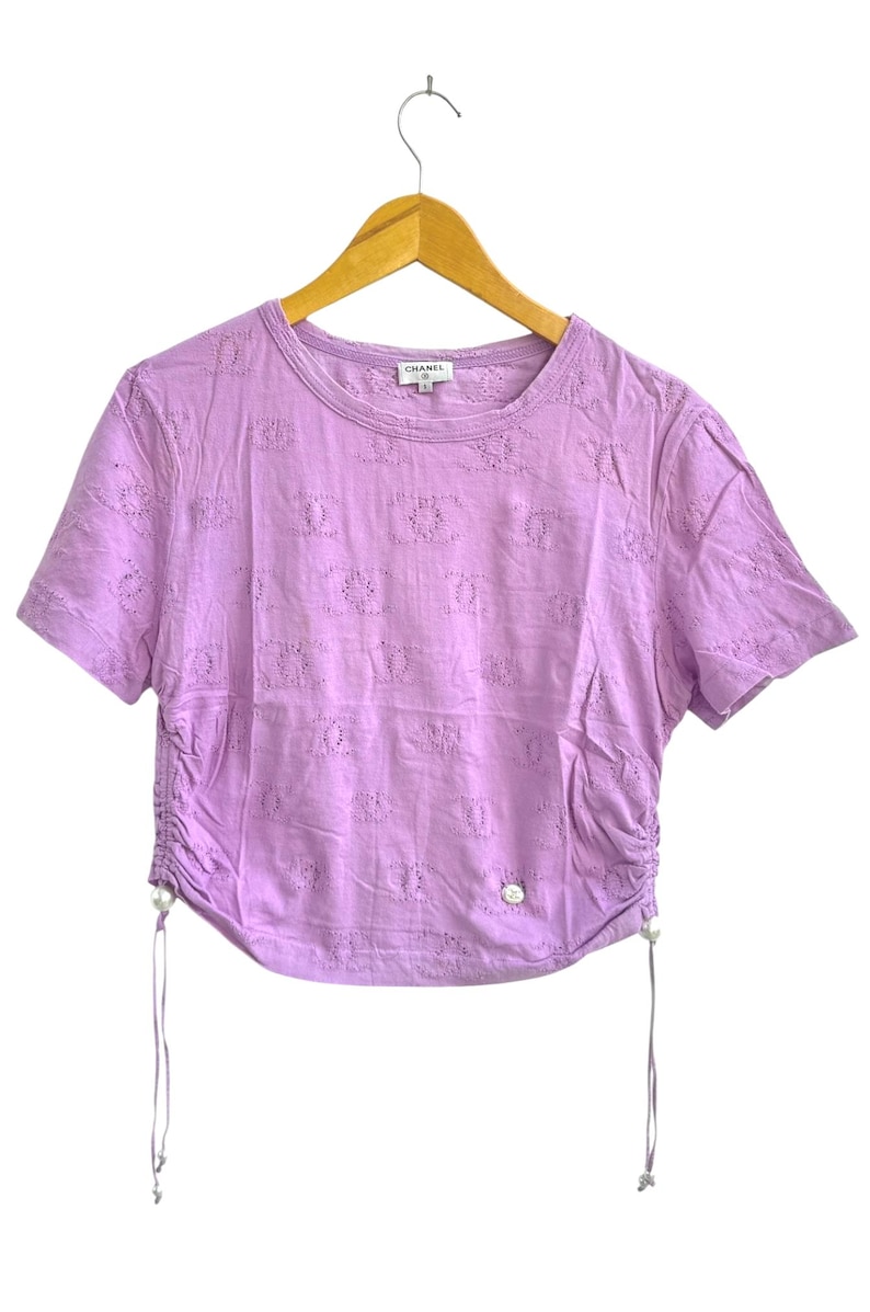 May include: A lavender short-sleeved t-shirt with a crew neck and a drawstring detail on the side. The shirt has a pattern of small, repeating shapes and a small logo near the neckline. The shirt is hanging on a wooden hanger.