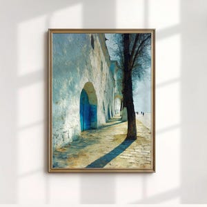 May include: Framed art print depicting a sunlit alleyway with white walls, arched doorways, and a blue door. A tall tree casts a long shadow across the stone path. The artwork has a textured, painterly style.