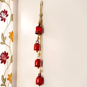 Peut inclure: Suspension murale décorative avec quatre cloches rouges suspendues à une corde naturelle. Les cloches sont cylindriques avec une base légèrement évasée. La corde est nouée à intervalles réguliers, et les cloches sont espacées uniformément. Fond blanc.