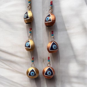 Könnte beinhalten: Zwei Stränge dekorativer Glocken. Jede Glocke hat eine metallische Gold- oder Kupferfarbe mit einem blauen Augendesign. Die Glocken sind mit Schnur und bunten Perlen aufgereiht. Der Hintergrund ist eine weiße, strukturierte Oberfläche.