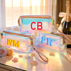 Op de afbeelding: Drie iriserende make-up tassen met gouden ritsen en polsbandjes. Elke tas heeft initialen in vette letters: "CB", "WM" en "IR". Een fles champagne met een strik staat op de achtergrond.