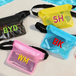 Op de afbeelding: Vier waterdichte heuptassen in zwart, geel, blauw en roze. Elke tas heeft een zwarte verstelbare riem en een doorzichtige voorkant met initialen in vette letters: BYD, SH, BK en WM. Zonnebrillen en een telefoon zijn zichtbaar in sommige tassen.
