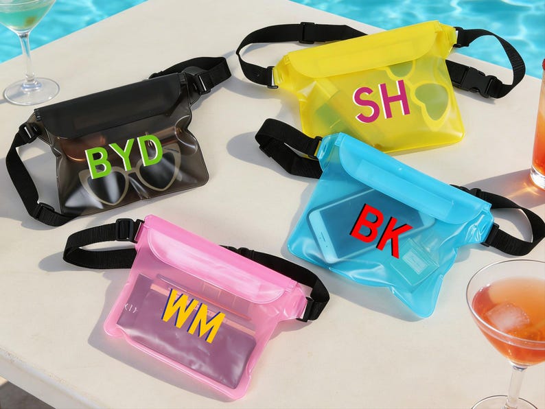 Op de afbeelding: Vier waterdichte heuptassen in zwart, geel, blauw en roze. Elke tas heeft een zwarte verstelbare riem en een doorzichtige voorkant met initialen in vette letters: BYD, SH, BK en WM. Zonnebrillen en een telefoon zijn zichtbaar in sommige tassen.