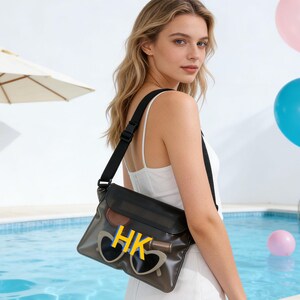Op de afbeelding: Een zwarte crossbody tas met een transparante voorkant, met de gele letters "HK" en een zonnebril. De tas heeft een zwarte band en wordt over de schouder gedragen. De achtergrond omvat een zwembad en kleurrijke ballonnen.