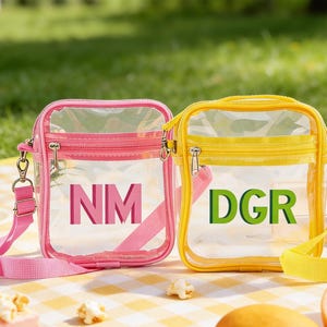 May include: Two clear crossbody bags, one pink and one yellow, with the letters "NM" and "DGR" respectively. Both bags have zip closures and adjustable straps. They are set on a yellow and white checked picnic blanket, with a green background.
