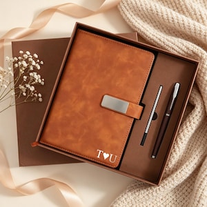 Pode incluir: Um caderno com aspeto de couro castanho com um fecho de metal prateado e o texto branco T coração U, apresentado numa caixa de presente castanha com uma caneta castanha escura a condizer e uma recarga de tinta prateada num tabuleiro.