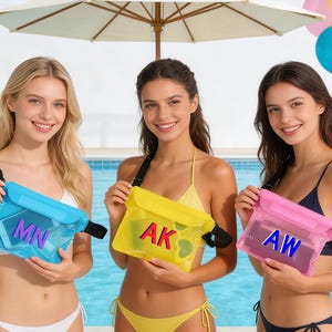 Könnte beinhalten: Drei Frauen halten wasserdichte Beutel in Blau, Gelb und Pink. Jeder Beutel hat einen schwarzen Riemen und eine transparente Vorderseite, die ein Telefon und die Initialen: MN, AK und AW anzeigt. Der Hintergrund umfasst einen Pool, Luftballons und einen Sonnenschirm.