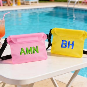 Op de afbeelding: Twee waterdichte crossbodytassen, &eacute;&eacute;n roze met groene letters "AMN" en de andere geel met blauwe letters "BH". Beide tassen hebben zwarte bandjes en staan op een witte tafel bij een zwembad.