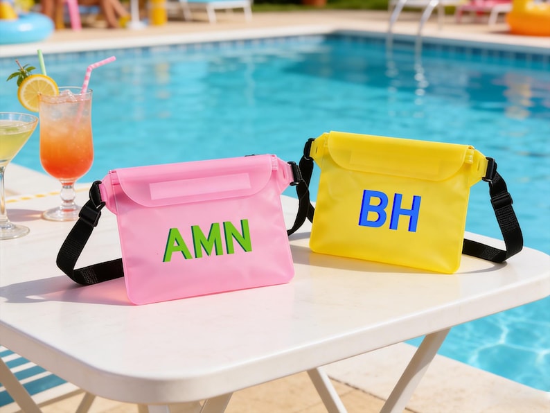 Op de afbeelding: Twee waterdichte crossbodytassen, &eacute;&eacute;n roze met groene letters "AMN" en de andere geel met blauwe letters "BH". Beide tassen hebben zwarte bandjes en staan op een witte tafel bij een zwembad.