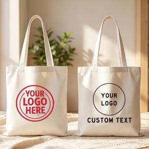 Può includere: Due borse tote in tela color bianco sporco con due tracolle. La borsa a sinistra presenta una grafica circolare rossa con il testo YOUR LOGO HERE. La borsa a destra mostra una grafica circolare nera con il testo YOUR LOGO e CUSTOM TEXT.