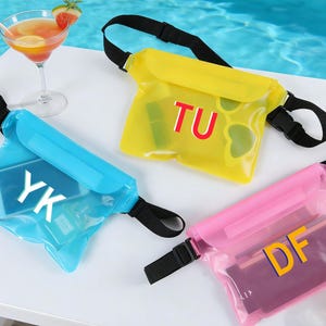 Op de afbeelding: Drie waterdichte heuptassen in blauw, geel en roze. Elke tas heeft een zwarte verstelbare riem en een gesp. De blauwe tas heeft een telefoon met de letters "YK". De gele tas heeft de letters "TU". De roze tas heeft de letters "DF".