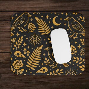 May include: Black mouse mat with a gold crow and botanical pattern. The design includes crows, ferns, stars, moons, and floral elements. A white computer mouse rests on the mat, next to a laptop.