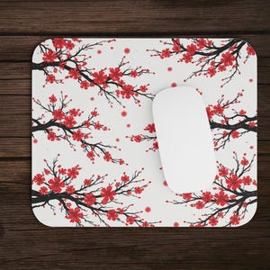 May include: A rectangular mouse mat with a white background featuring a repeating pattern of red cherry blossom branches. A white computer mouse rests on the mat. A laptop is partially visible.
