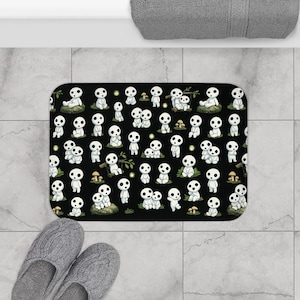 Puede incluir: Una alfombra de baño rectangular negra con un patrón repetido de espíritus del bosque blancos de dibujos animados. Los espíritus tienen ojos grandes y redondos y se representan en varias poses. La alfombra está colocada sobre un suelo embaldosado, con zapatillas grises visibles.