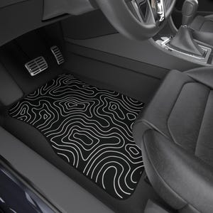May include: Black car floor mat featuring a white topographic map pattern. The mat is rectangular with a curved edge, designed for the driver's side footwell. The mat is positioned in a car, near the pedals and the driver's seat.