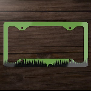 May include: A green license plate frame with a black and gray forest scene at the bottom. The forest design includes silhouettes of trees and a small cabin. The frame has a rectangular shape with rounded corners.